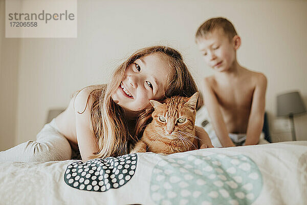 Lächelndes Mädchen  das sich an eine Katze lehnt  während der Bruder im Hintergrund im Schlafzimmer zu Hause ist