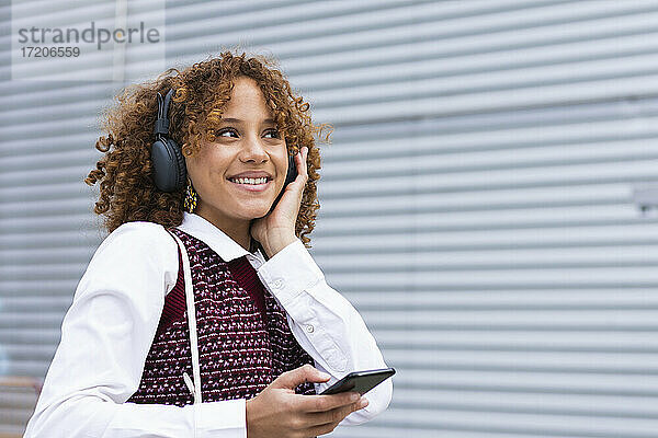 Lächelnder weiblicher Teenager mit Mobiltelefon  der über Kopfhörer an der Wand Musik hört