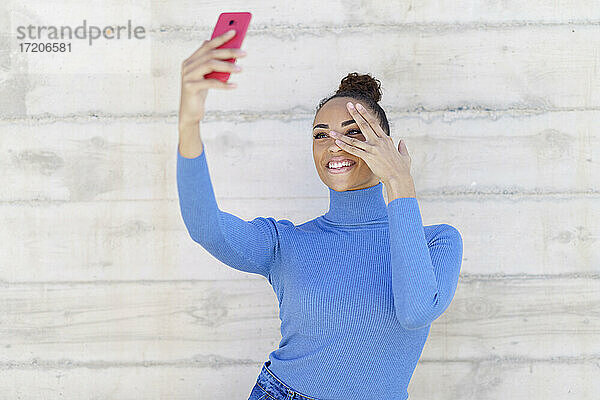 Lächelnde Frau gestikuliert  während sie ein Selfie durch ein an der Wand stehendes Mobiltelefon macht