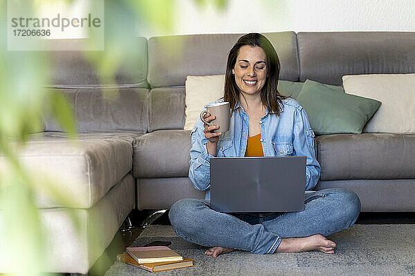 Lächelnde Frau trinkt Kaffee und benutzt einen Laptop im Wohnzimmer