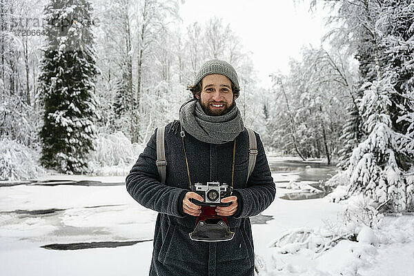 Junger Mann mit Kamera  der lächelnd im verschneiten Wald steht