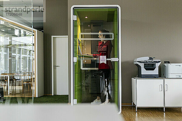 Unternehmerin  die in einer schalldichten Kabine im Büro mit ihrem Smartphone telefoniert