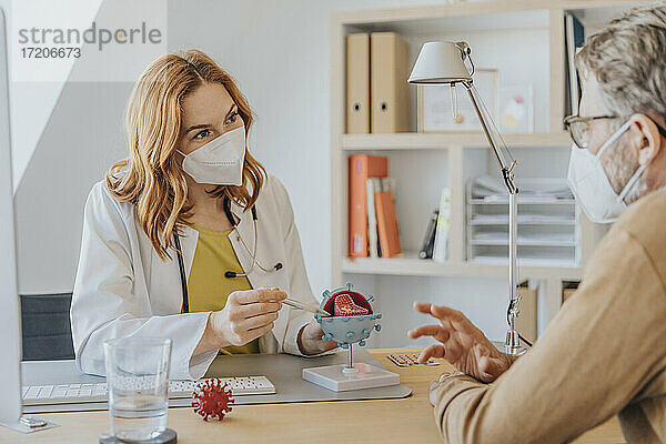 Ärztin mit Gesichtsschutzmaske erklärt einem Patienten im Büro sitzend ein künstliches Coronavirus-Modell