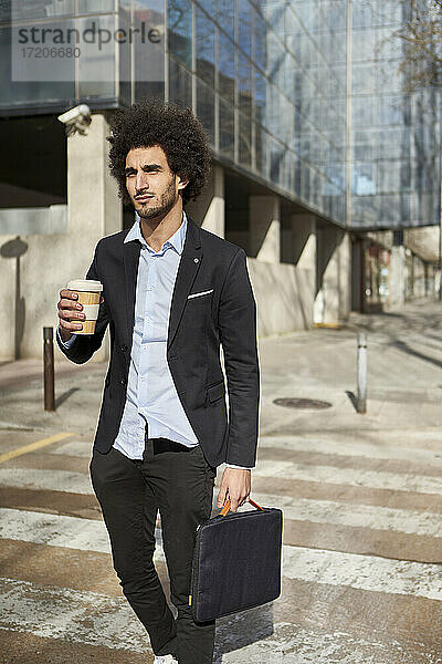 Geschäftsmann mit Laptoptasche und Kaffeetasse beim Überqueren der Straße an einem sonnigen Tag