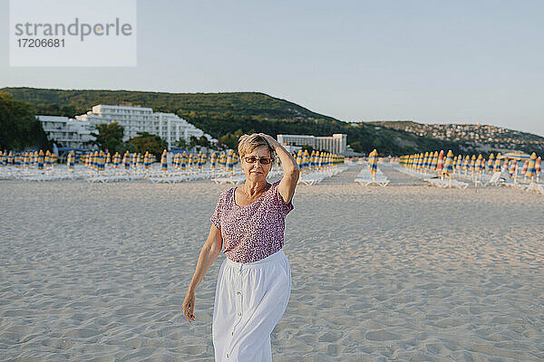 Ältere Frau mit Hand in den Haaren steht gegen einen Hügel am Strand