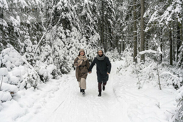 Lächelndes Paar  das sich an den Händen hält  während es auf Schnee im Wald läuft