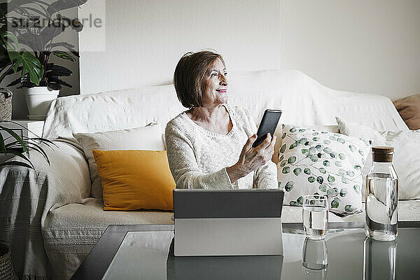 Lächelnde ältere Frau mit Laptop und Handy in der Hand  während sie zu Hause auf dem Sofa sitzt