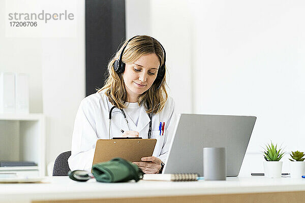 Medizinische Mitarbeiterin schreibt auf einem Klemmbrett  während sie eine Online-Konsultation über einen Laptop in einer medizinischen Klinik durchführt