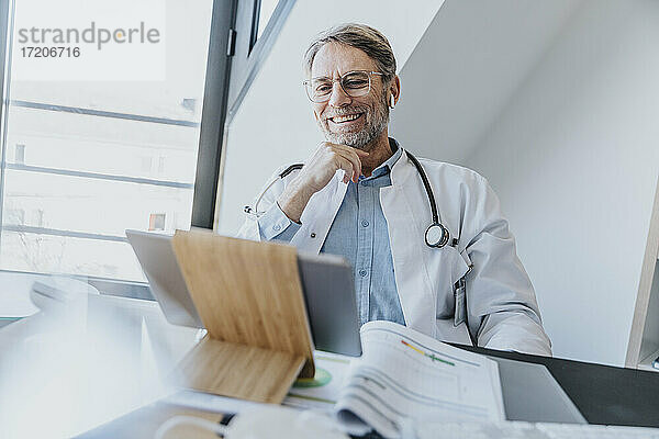Älterer Arzt lächelnd bei der Verwendung eines digitalen Tablets in der Arztpraxis