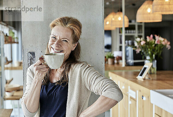 Glückliche reife Frau mit Hand auf der Hüfte beim Kaffee trinken gegen Säule zu Hause