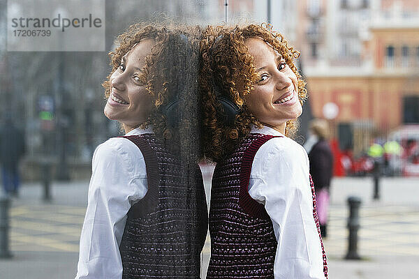 Lächelndes Teenager-Mädchen mit Kopfhörern  das sich an eine Glaswand lehnt