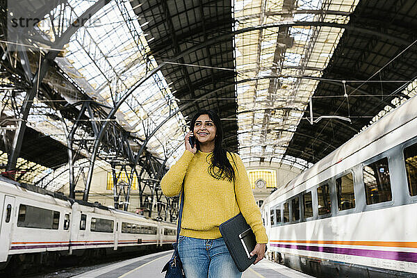 Lächelnde Frau mit Laptoptasche  die wegschaut  während sie auf dem Bahnsteig mit ihrem Smartphone telefoniert