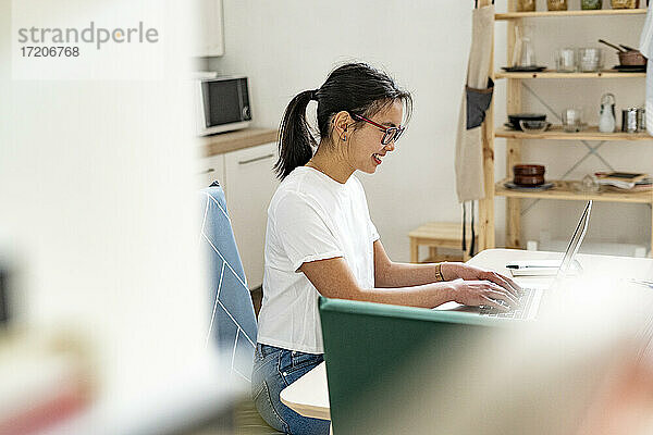 Lächelnde Frau arbeitet am Laptop  während sie in der Küche zu Hause sitzt
