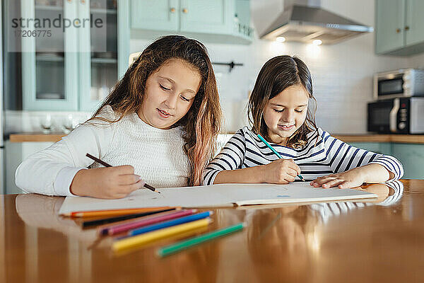 Schwestern malen mit Buntstiften auf Papier  während sie am Esstisch in der Küche sitzen