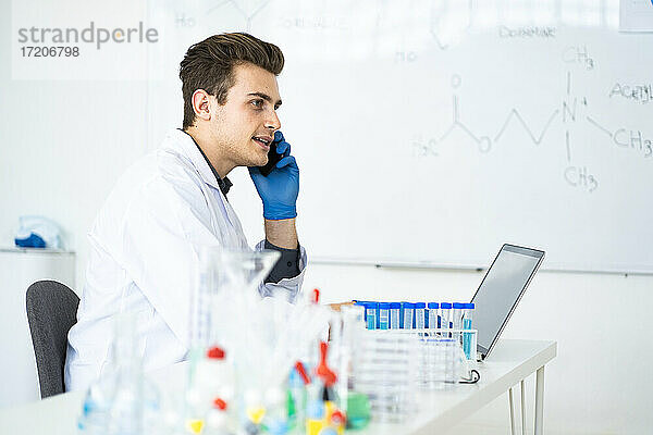 Männlicher Wissenschaftler  der am Schreibtisch sitzend in einem Labor mit einem Mobiltelefon spricht