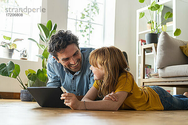 Lächelnde Tochter mit digitalem Tablet  die ihren Vater anschaut  während sie zu Hause im Wohnzimmer auf dem Boden liegt