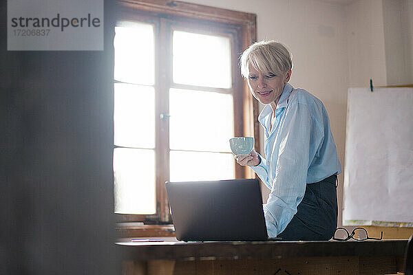 Lächelnde Geschäftsfrau  die eine Kaffeetasse hält  während sie einen Laptop auf dem Schreibtisch im Büro zu Hause benutzt
