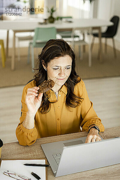 Geschäftsfrau  die einen Keks isst  während sie einen Laptop am Schreibtisch im Heimbüro benutzt