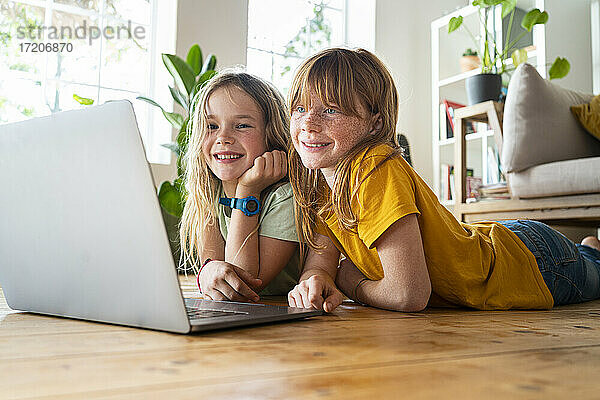 Lächelnde Freundinnen schauen auf einen Laptop  während sie zu Hause im Wohnzimmer auf dem Boden liegen