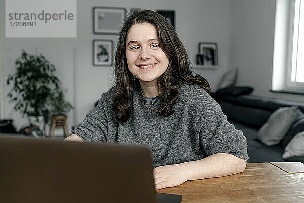 Lächelnde Geschäftsfrau sitzt mit Laptop am Tisch im Wohnzimmer