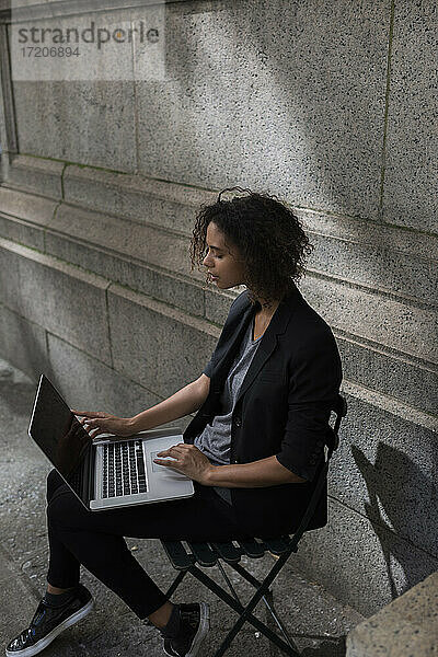 Afro-Unternehmerin  die einen Laptop benutzt und auf einem Stuhl vor einer Wand sitzt