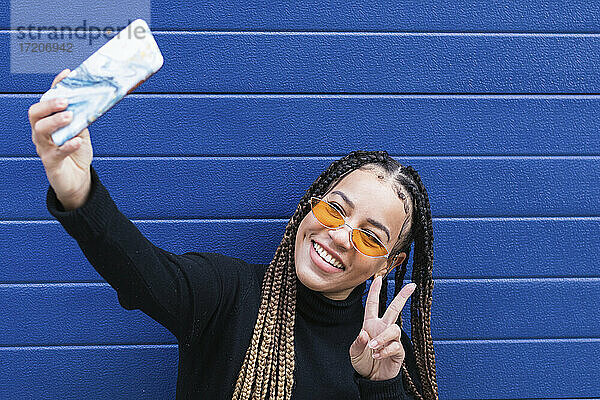 Lächelnde Frau  die ein Selfie mit ihrem Mobiltelefon macht  während sie an einer blauen Wand steht