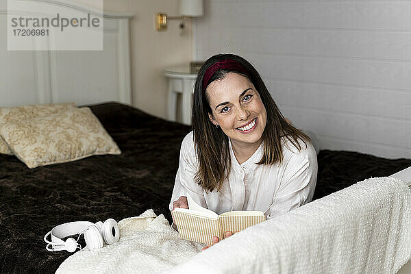Lächelnde Frau mit Buch auf dem Bett liegend zu Hause