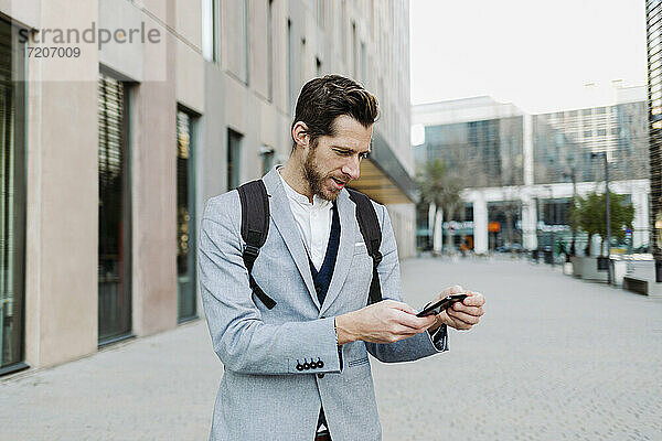 Männlicher Unternehmer mit Rucksack  der ein Mobiltelefon benutzt  während er an einem Gebäude steht