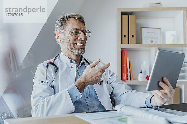 Lächelnder Arzt  der über ein digitales Tablet einen Videoanruf führt  während er in einer Klinik sitzt