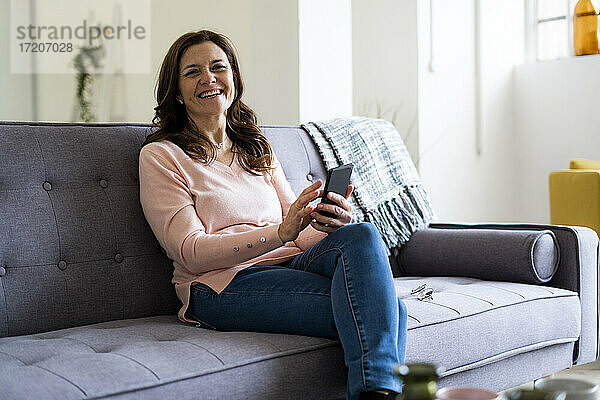 Glückliche reife Frau  die ein Mobiltelefon hält  während sie auf dem Sofa im Wohnzimmer sitzt