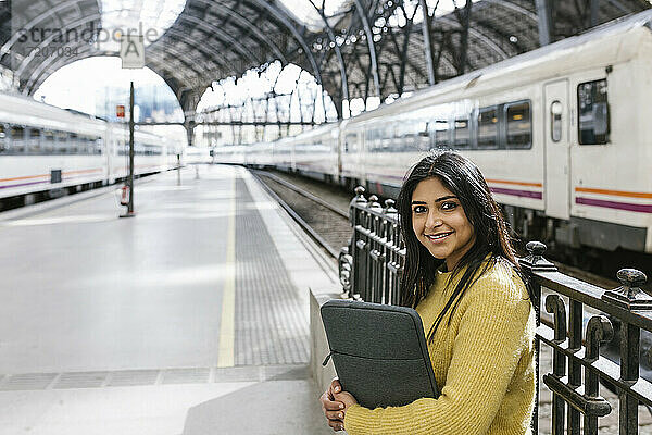 Lächelnde mittelalte Frau mit Laptoptasche am Geländer auf dem Bahnsteig