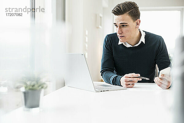 Junger männlicher Berufstätiger mit Smartphone  der im Büro zu Hause auf seinen Laptop schaut