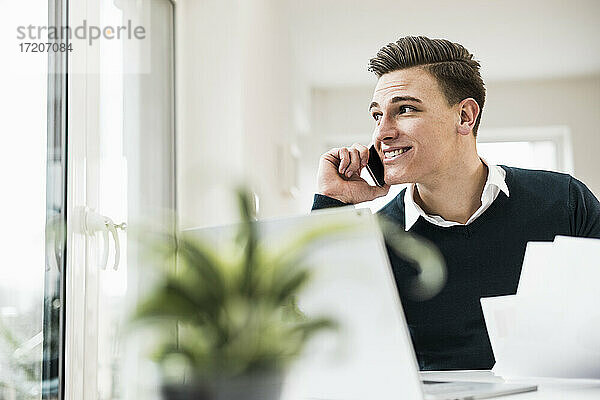 Lächelnder männlicher Unternehmer  der am Telefon spricht und durch ein Fenster im Büro zu Hause schaut