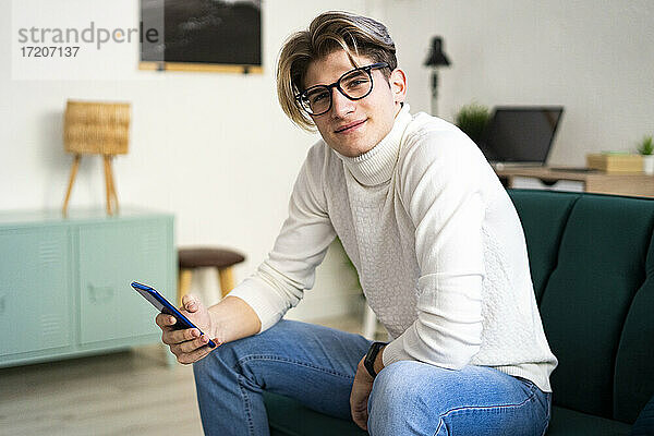 Lächelnder junger Mann mit Smartphone auf dem Sofa im Wohnzimmer sitzend
