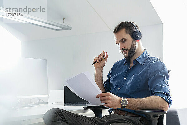 Junger Geschäftsmann mit Kopfhörern  der im Büro sitzt und Papier prüft