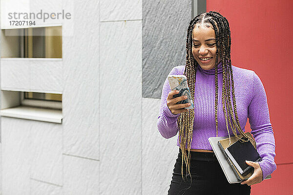 Lächelnde Studentin  die Bücher in der Hand hält und ein Mobiltelefon benutzt  während sie an der Wand steht