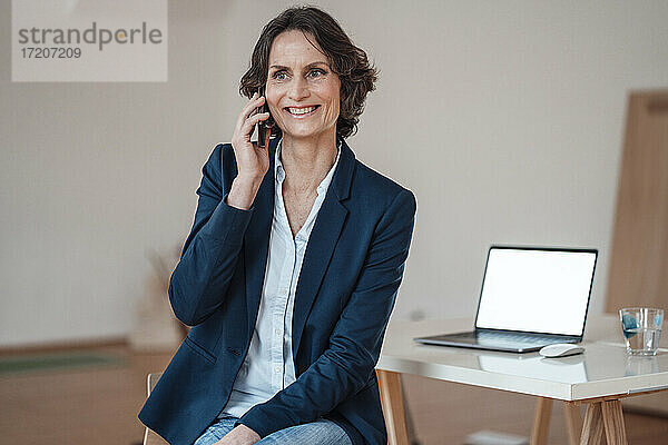Lächelnde Unternehmerin  die mit ihrem Smartphone telefoniert  während sie mit ihrem Laptop auf dem Tisch im Heimbüro sitzt