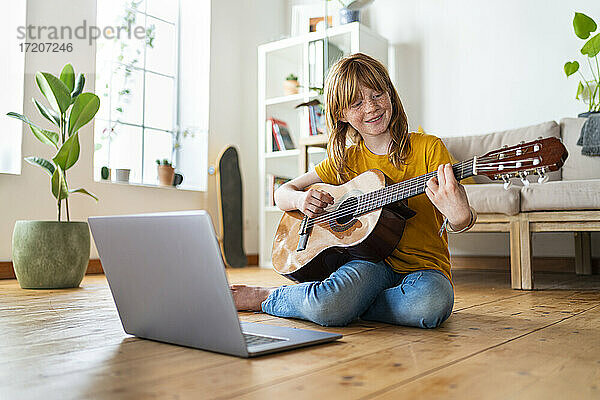 Lächelndes rothaariges Mädchen  das Gitarre spielt  während es zu Hause mit dem Laptop e-learning betreibt