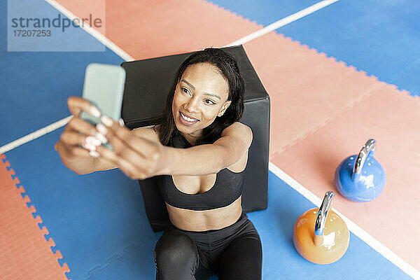 Fröhliche junge Frau  die ein Selfie macht  während sie sich im Fitnessstudio auf eine Box stützt