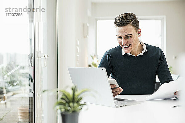 Fröhlicher Geschäftsmann  der eine SMS auf seinem Smartphone schreibt  während er im Home Office sitzt