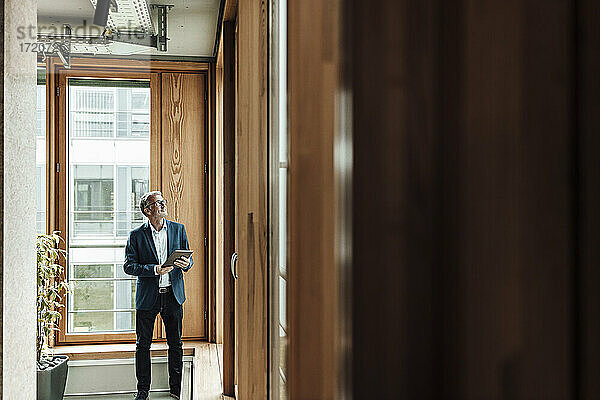 Älterer Geschäftsmann mit digitalem Tablet  der im Bürokorridor steht und nach oben schaut