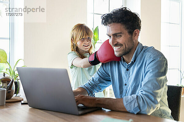 Lächelndes rothaariges Mädchen mit Boxhandschuhen  das seinen Vater schlägt  der zu Hause am Laptop arbeitet