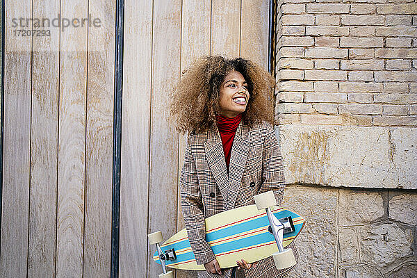 Fröhliche junge Frau mit Skateboard an der Wand stehend