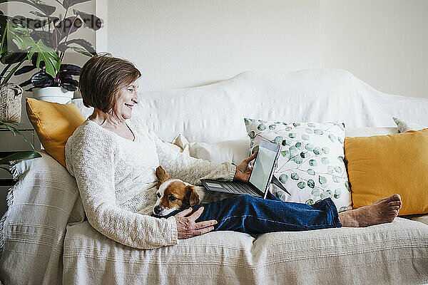 Ältere Frau benutzt einen Laptop  während sie mit ihrem Jack Russell Terrier zu Hause auf dem Sofa sitzt