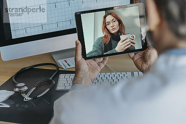 Männlicher Arzt im Gespräch mit einem Patienten per Videoanruf über ein digitales Tablet  während er in einer Arztpraxis sitzt
