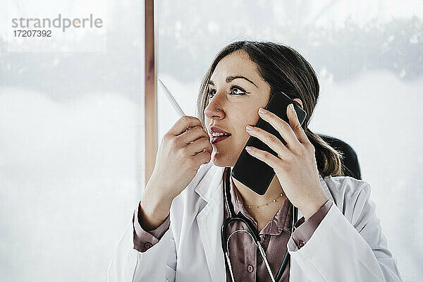 Nachdenkliche Ärztin  die wegschaut  während sie mit einem Mobiltelefon spricht