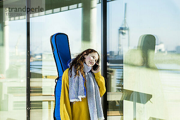 Lächelndes Mädchen  das eine Gitarre trägt  während es mit einem Mobiltelefon an einem Glas spricht