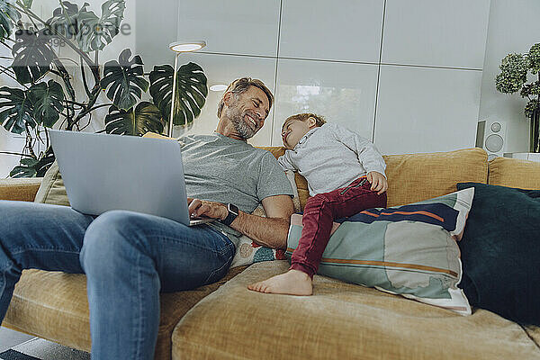 Glücklicher Vater mit Laptop und Sohn auf dem Sofa im Wohnzimmer