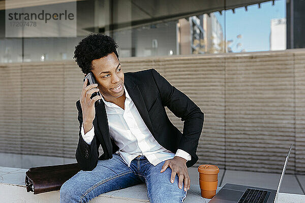 Lächelnder männlicher Geschäftsmann  der auf einer Stützmauer sitzend mit einem Smartphone spricht und dabei wegschaut