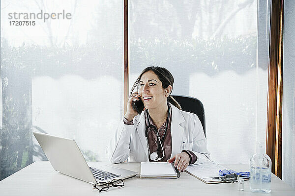 Lächelnde Ärztin  die wegschaut  während sie mit einem Smartphone in einer medizinischen Klinik spricht
